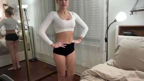 Media: A video of a fit, light-skinned woman with blonde hair in a white crop top and black shorts, standing confidently in a bedroom with a bed, mirror, and lamp.