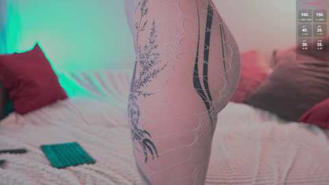 Media: Video of a woman's lower back and buttocks in white lace lingerie, showing intricate lace patterns and black tattoos. She stands on a bed with red pillows and a green blanket, bathed in soft, pastel-colored lighting.