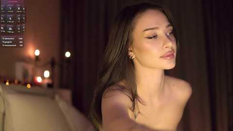 Media: Video of a slender, topless woman with long brown hair, fair skin, and small breasts, gazing thoughtfully. Dimly lit room with a television screen displaying a digital clock and blurred background.