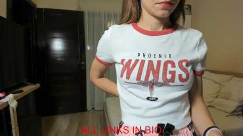 Media: Video of a young woman with light skin, medium-length brown hair, wearing a tight white t-shirt with red trim and \"Phoenix Wings\" logo, standing in a modern, beige-toned living room with a TV, couch, and door.