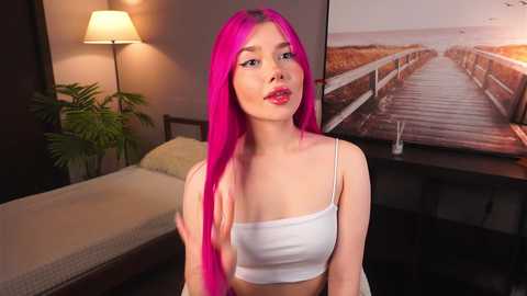 Media: A video of a young woman with vibrant pink hair and fair skin, wearing a white crop top, standing in a modern bedroom with a potted plant, lamp, and framed beach scene on the wall.