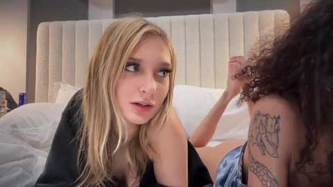 Media: Video of two young women in a bed, one with long blonde hair, wearing a black top, and the other with curly brown hair, both in casual attire.