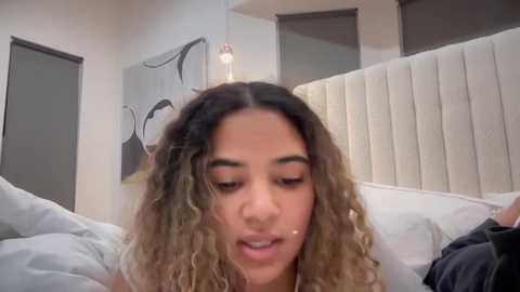 Media: Video of a young woman with light brown skin, curly hair, and makeup, lying on a bed with white pillows and a beige headboard. The background features minimalist decor with abstract art and a modern, clean aesthetic.