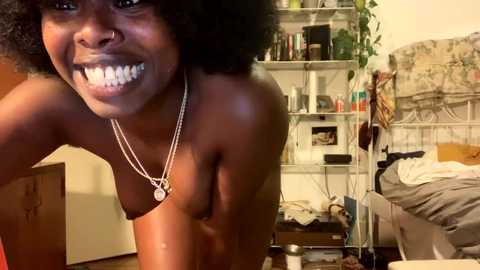 Media: Video of a topless Black woman with an afro, smiling broadly, in a cluttered bedroom with bookshelves, a bed, and a TV.