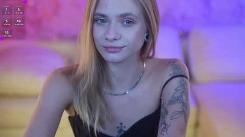 Media: Video of a slim, young Caucasian woman with long, straight blonde hair, blue eyes, and fair skin, wearing black strapless top, and tattoos on her shoulder. Background features purple and yellow hues.