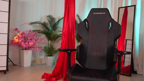 Media: Video of a black gaming chair with red accents, surrounded by lush green plants and pink orchids, with red drapes and a white partition in a minimalist, modern room.