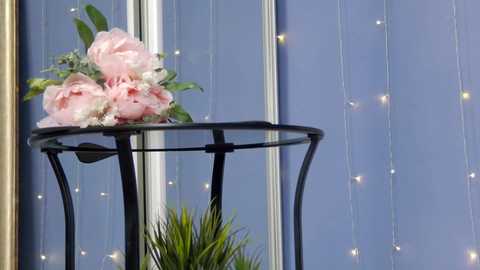 Media: A video featuring a black metal garden stool with a bouquet of soft pink peonies and green leaves, set against a blue wall adorned with fairy lights, with a small potted plant in the foreground.