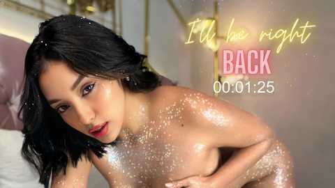 Media: Video of a nude, attractive, light-skinned Asian woman with shoulder-length black hair, glistening with glitter, posing seductively on a bed. Background features a luxurious, golden bedroom setting with soft lighting. Text reads \"I'm the night's back\" and \"00:01:25.\