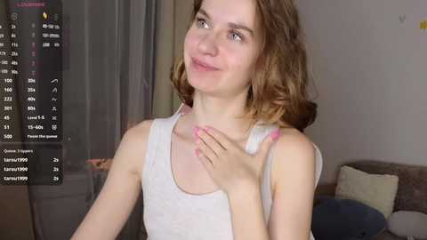 Media: Video of a young woman with fair skin and wavy brown hair, wearing a white tank top, smiling and touching her chest, indoors with a sofa and curtains in the background.