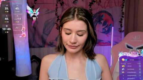 Media: Video of a young woman with shoulder-length brown hair in a light blue halter top, sitting in a gaming chair with a monitor showing a \"PewDiePie\" stream. Background features a colorful, abstract mural and fairy-themed decor.