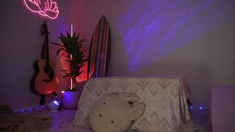 Media: Video of a cozy room with a white tablecloth-covered table, a plush bear, guitar, potted plant, and colorful neon lights on a gray wall.