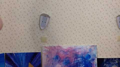 Media: Video of a classroom wall with white, textured wallpaper. A large, colorful abstract painting with blue and pink hues dominates the lower portion. Above, a gray, childlike face cutout is mounted. The background is a mix of blue and purple abstract art.