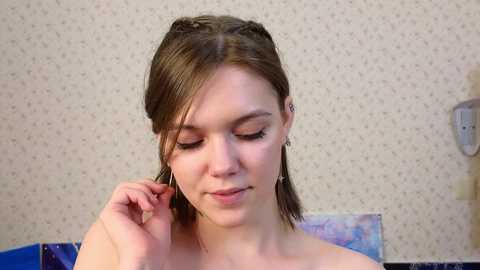 Media: Video of a young Caucasian woman with light skin, brown hair styled in two braids, and light makeup. She's topless, adjusting her hair, in a room with a beige patterned wallpaper and a blue couch in the background.