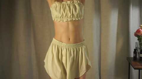 Media: Video of a slender, fair-skinned woman in a cream-colored strapless crop top and matching shorts, standing against a beige curtain backdrop with a vase of flowers on a wooden table to the right.