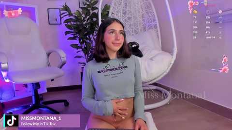 Media: Video of a young woman with light skin and shoulder-length dark hair, wearing a cropped light blue top, sitting in a modern bedroom with white furniture, plants, and a white hanging chair.