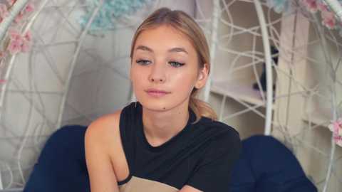 Media: Video of a fair-skinned young woman with blonde hair in a black, off-shoulder top, sitting in a white, flower-adorned hanging chair. Background shows a blurred, pastel-colored room.