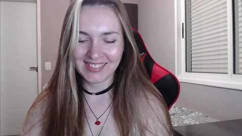 Media: Video of a smiling young woman with long, straight brown hair, wearing a black choker necklace and red pendant, seated indoors with a gaming chair and window in the background.