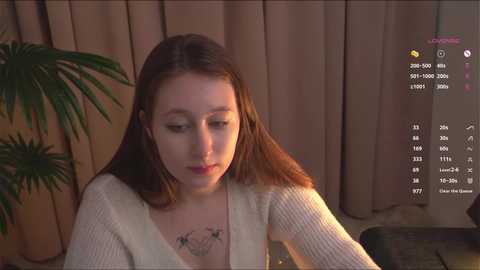 Media: Video of a young woman with light skin and brown hair, wearing a white knitted cardigan, seated in a dimly lit room with beige curtains and a potted plant.