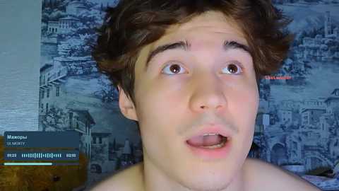 Media: Video of a young, shirtless man with curly brown hair and wide eyes, mouth open in surprise. Background features a blue and white mural of a European cityscape.