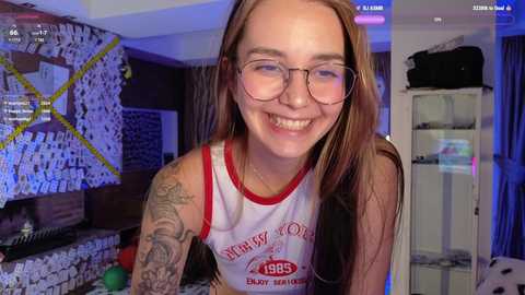 Media: Video of a young woman with fair skin, long brown hair, wearing glasses and a white tank top with red trim, smiling in a dimly lit room with posters and a bookshelf.