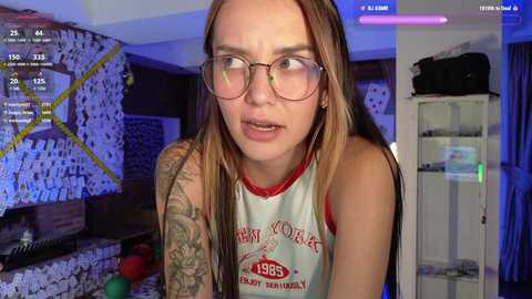 Media: Video of a young woman with long brown hair, glasses, and tattoos, wearing a white tank top with \"1985\" and \"Hawaii\" text, in a dimly lit room with blue and purple lighting.