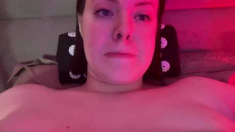 Media: Video of a light-skinned woman with short dark hair, lying topless on a bed, illuminated by pink and red lights. She has circular earrings with white patterns.