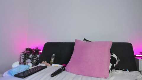 Video of a minimalist bedroom with a black headboard, a pink pillow, and a TV remote on a white bedspread. The walls are plain white, and pink ambient lighting creates a soft, calming atmosphere.