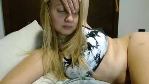 Media: Video of a young, fair-skinned, blonde woman with shoulder-length hair, wearing a tie-dye crop top, lying on a bed with white sheets, covering her face with her hand.