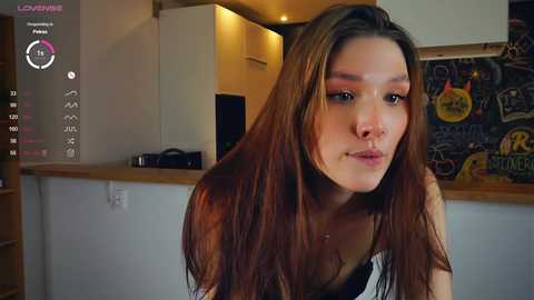 Media: Video of a young woman with long, straight brown hair and fair skin, wearing a black top, in a modern kitchen with wooden cabinets and a colorful, cluttered wall.