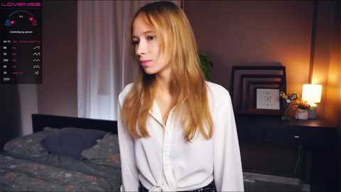 Media: A video of a young woman with long blonde hair, wearing a white button-up shirt, standing in a dimly-lit bedroom.