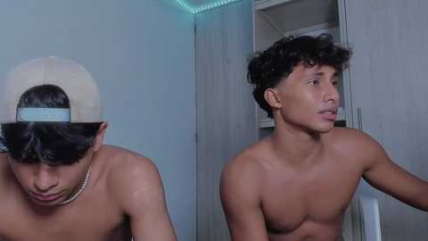 Media: Video of two shirtless young men in a dimly lit room. One wears a backward cap, the other curly hair; both appear surprised, with a closet in the background.