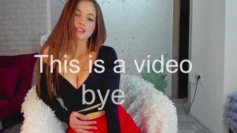 Media: Video of a light-skinned woman with long, straight brown hair, wearing a black crop top and red skirt, sitting on a white fur blanket in a modern living room. Text overlay: \"This is a video by bye.\