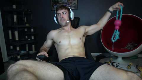 Media: Video of a shirtless, muscular man with light skin, short brown hair, and blue eyes, wearing headphones, sitting in a gaming chair, holding a game controller, and a pink and turquoise toy in a dark room.