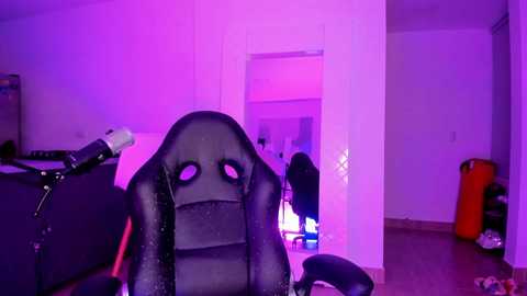 Media: A video of a dimly lit, minimalist bedroom with purple lighting. A black gaming chair with a large, padded headrest dominates the foreground. A doorway reveals a dark room with an open door. The room has a cluttered feel with a red and white air pump on the right.