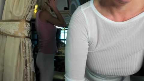 Media: Video of a woman in a white ribbed top, partially obscured by a blonde woman in a pink tank top, standing in a dimly lit, ornately decorated room.