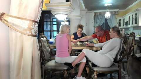 Media: Video of a middle-aged woman with blonde hair, wearing a pink tank top and gray pants, seated at a wooden table with a bald man in a red shirt, a woman with brown hair in a black top, and a man in glasses in a white sweater.