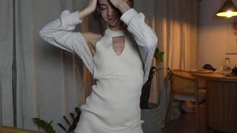 Media: Video of a slender woman with light skin, wearing a cream-colored, ribbed knit dress with a keyhole cutout, standing in a dimly lit room with beige curtains, a wooden table, and a green plant.