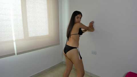 Media: Video of a young Asian woman with long black hair, wearing black lingerie, standing against a white wall in a minimalist room with a window.