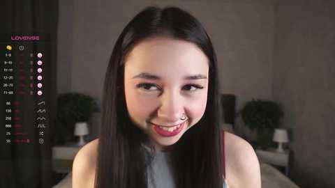 Media: A video of a young Asian woman with long black hair, fair skin, and red lipstick, smiling playfully, wearing a white tank top. The background features a dimly lit bedroom with a bed, plants, and a lamp.