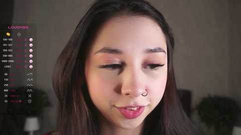 Media: Video of an Asian woman with straight black hair and fair skin, wearing a septum ring and subtle makeup, in a dimly lit room.