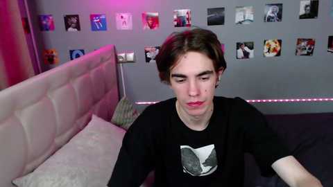 Media: A video of a young man with light skin and dark hair, wearing a black shirt with a white graphic, sitting on a bed in a dimly lit room with pink lighting and posters on the wall.