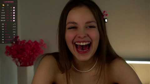 Media: Video of a young, light-skinned woman with straight brown hair, wearing a pearl necklace, laughing joyfully with open mouth. Background features a blurred, beige wall with a pink flower arrangement on the left and a digital clock on the top left.