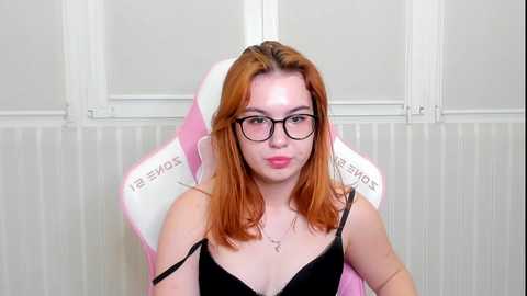 Media: Video of a young Caucasian woman with fair skin, shoulder-length red hair, and glasses, sitting in a pink gaming chair, wearing a black camisole, against a white-paneled wall.