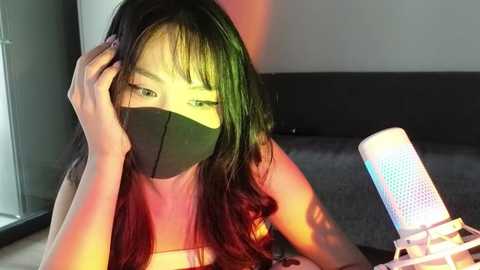 Media: Video of an Asian woman with shoulder-length, green and red hair, wearing a black face mask and a white top, sitting with a white, light-up microphone on a grey carpeted floor.