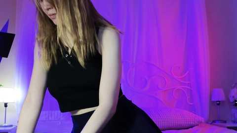 Media: Video of a young woman with fair skin and long blonde hair, wearing a black sleeveless top and high-waisted skirt, leaning forward, in a dimly lit bedroom with purple and white lights.