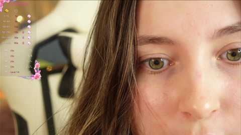 Media: A close-up video of a young woman with light brown hair, green eyes, and a neutral expression. In the background, a blurred mannequin and a calendar are visible.