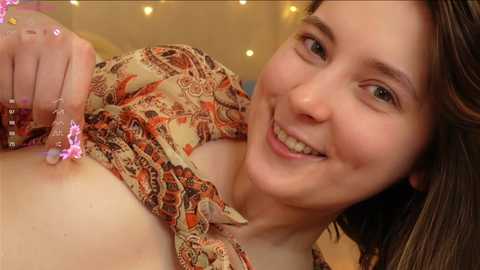 Media: Video of a smiling young woman with fair skin and brown hair, wearing a patterned blouse, revealing her left breast, with a pink glittery bracelet on her wrist. Background shows warm, fuzzy lights.