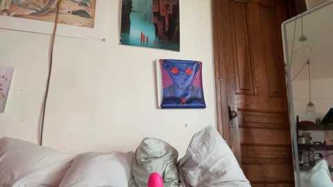 Media: Video of a messy, lived-in bedroom with white walls, a wooden door, a mirror, and abstract art, featuring a blue face with red eyes and a pink vibrator on a bed.