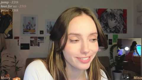 Media: Video of a young woman with light skin, brown hair, and a nose ring, smiling in a cozy room with colorful art and plants.