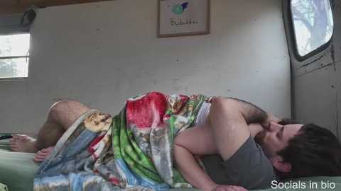 Media: Video of a man lying on a bed in a small, dimly lit room, covered by a colorful blanket. The room has a plain white wall with a framed logo.